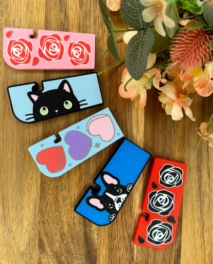 A selection of five colorful Tab Buddy XLs with cat, rose, dog, and heart designs on a wooden surface with flowers.