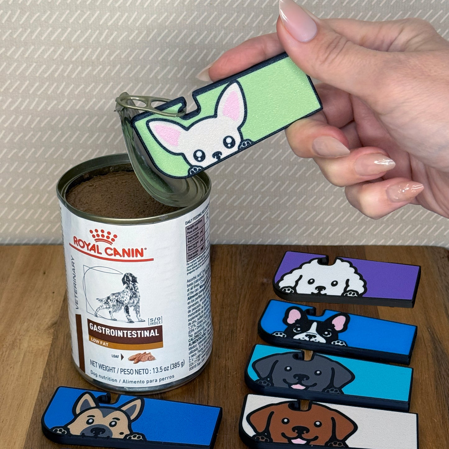 Hand using a Tab Buddy XL with a Chihuahua-inspired design to open a can of dog food against a beige background. Additional Tab Buddy XLs with related dog desigs rest on a wooden surface.