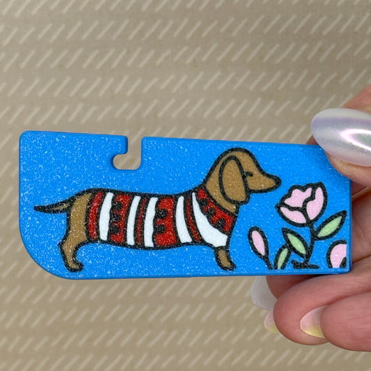 A sky blue Tab Buddy XL with image of a light brown dachshund dog smelling pink flowers is held in a hand with beige background.
