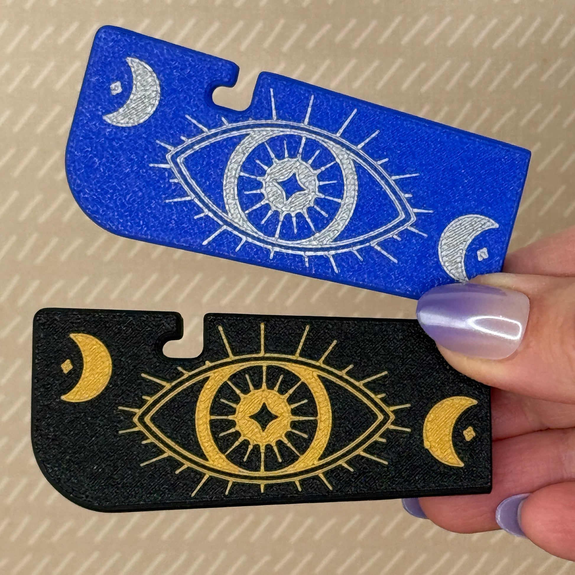 Two Tab Buddy XLs with eye and moon designs, one blue and one black, held by a hand.
