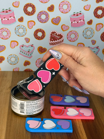 Hand holding a black Tab Buddy XL with heart designs against a colorful wallpaper background. Three more color variations of the heart-designed Tab Buddy XL rest on a wooden surface.