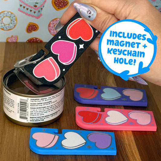 Hand holding a black Tab Buddy XL with heart designs against a colorful wallpaper background. Three more color variations of the heart-designed Tab Buddy XL rest on a wooden surface. Text box reads, "Includes magnet + keychain hole!"