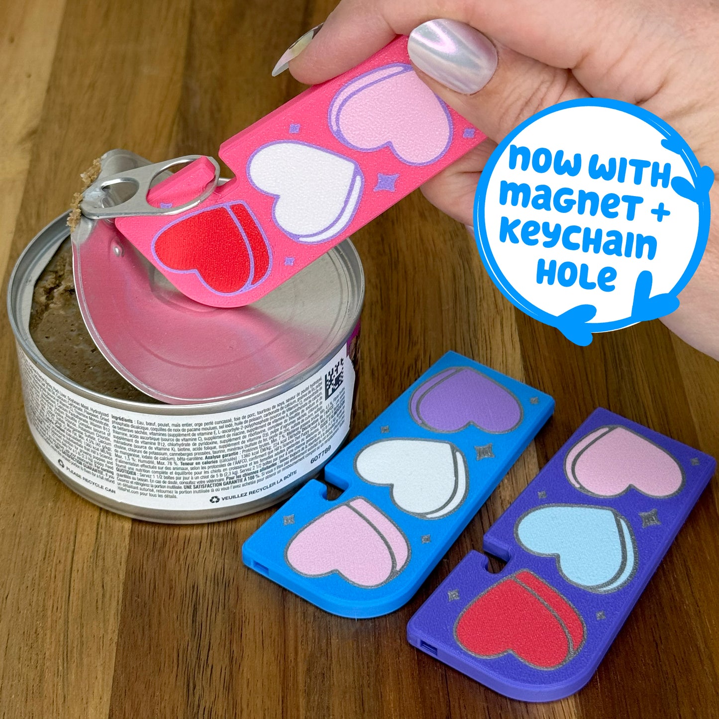 Pink Tab Buddy XL with Candy Hearts design is used to open a can of food. Different color variations of the same hearts design Tab Buddy XLs rest on a wooden surface. Each on has a magnet and keychain hole.