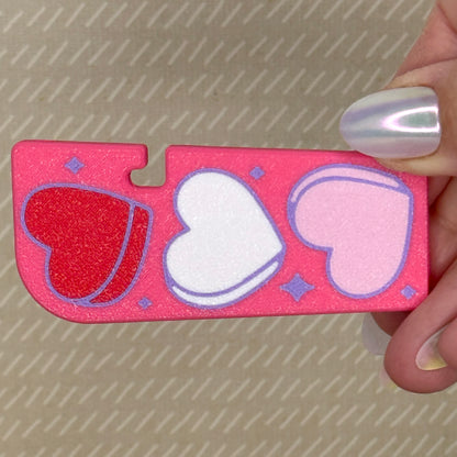 Bright pink Tab Buddy XL with light pink, white, and red heart shapes held by a hand against a textured background
