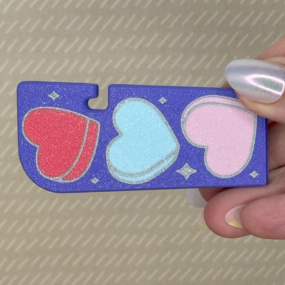 Purple Tab Buddy XL with pink, blue, and red heart shapes held by a hand against a textured background