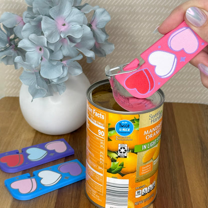 Pink Tab Buddy XL with Candy Hearts design is used to open a can of food. Different color variations of the same hearts design Tab Buddy XLs rest on a wooden surface. Each on has a magnet and keychain hole.