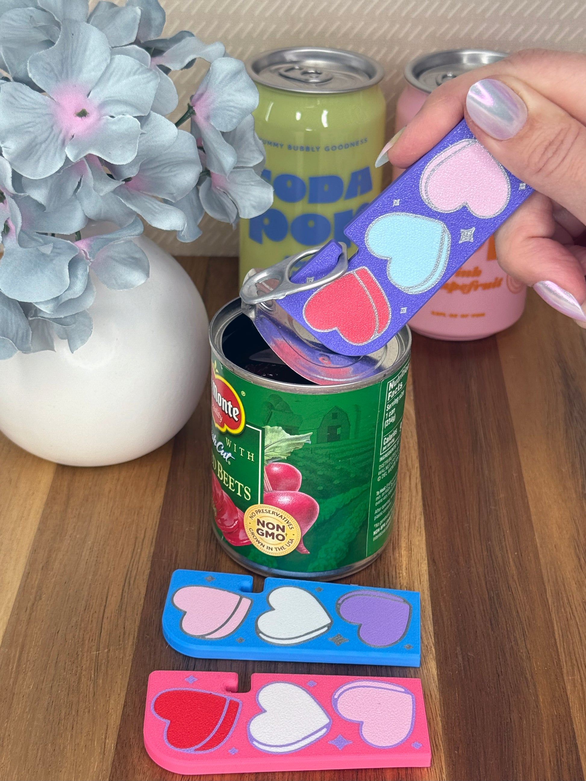 Purple Tab Buddy XL with Candy Hearts design is used to open a can of food. Different color variations of the same hearts design Tab Buddy XLs rest on a wooden surface. Each on has a magnet and keychain hole.