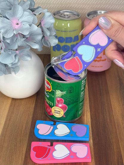 Purple Tab Buddy XL with Candy Hearts design is used to open a can of food. Different color variations of the same hearts design Tab Buddy XLs rest on a wooden surface. Each on has a magnet and keychain hole.