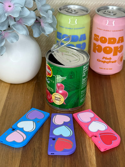 Three Tab Buddy XLs with Candy Hearts designs rest on a wooden surface in front of a can of opened beets. Soda cans and flowers are in the background.