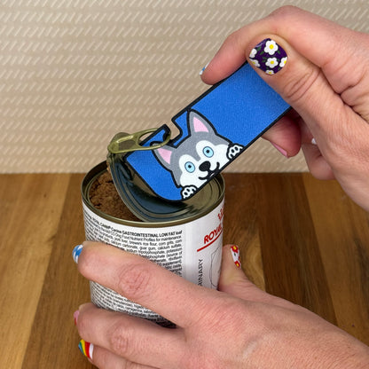 Hand using a Tab Buddy XL with a Husky dog design to open a can of dog food on a wooden surface.