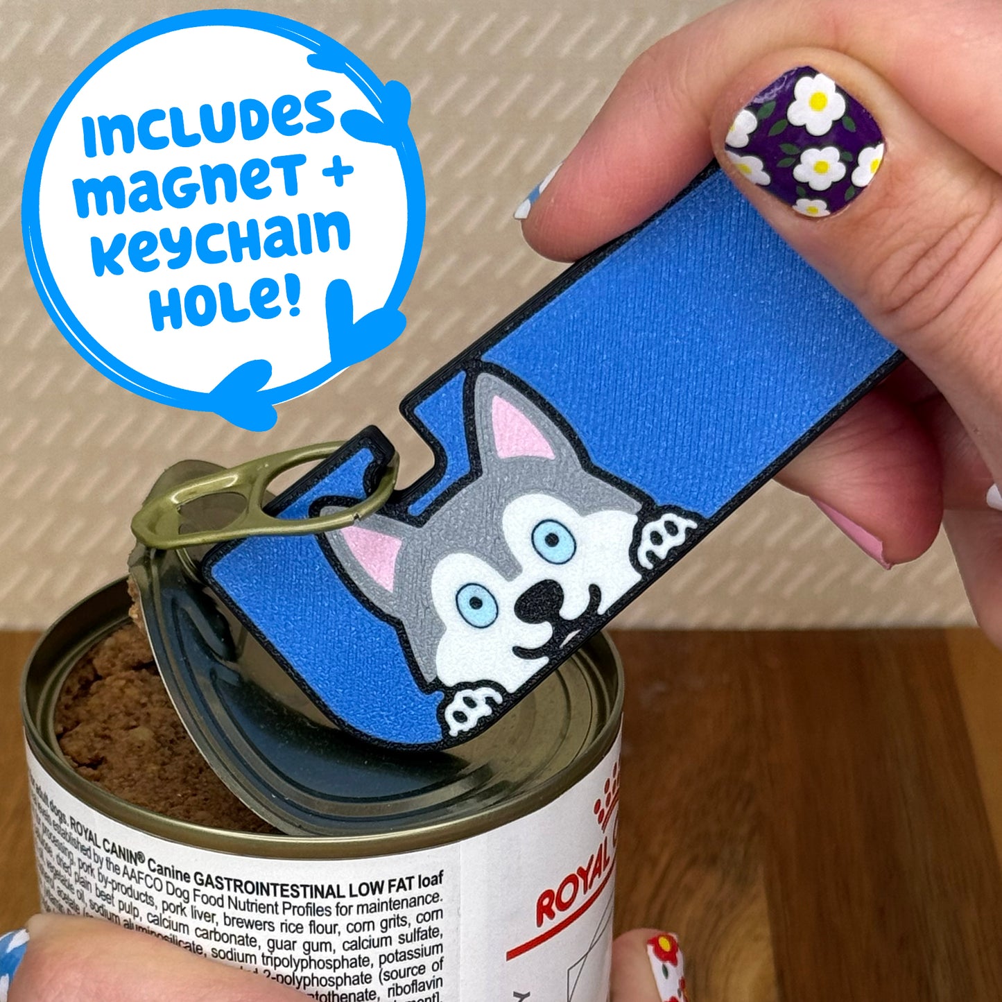 Hand using a Tab Buddy XL with a Husky dog design to open a can of dog food on a wooden surface. Text bubble reads, "Includes magnet + keychain strap!"
