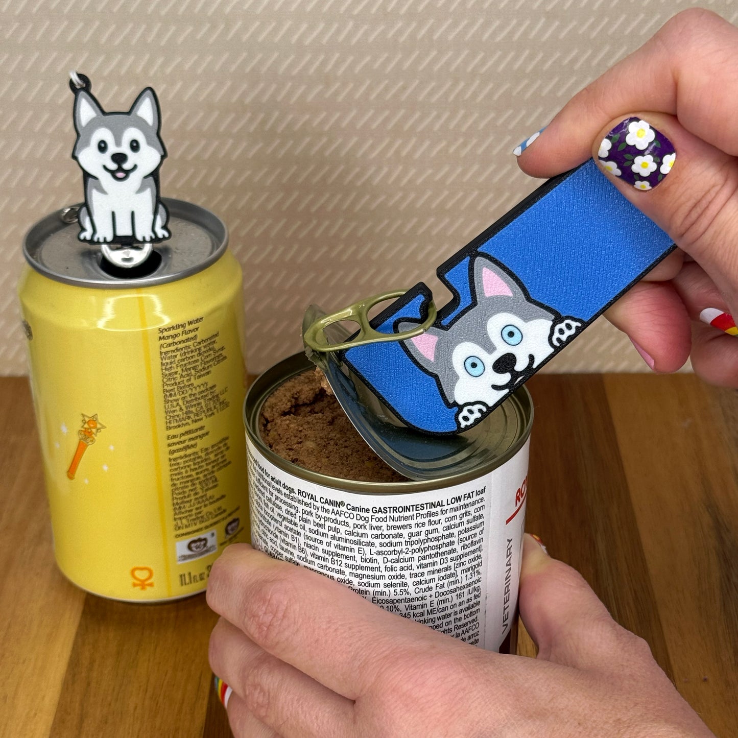 A person opens a can of dog food with a grey Husky dog-design Tab Buddy XL. In the background, a matching soda-sized Tab Buddy rests atop the tab of a can of sparkling water.