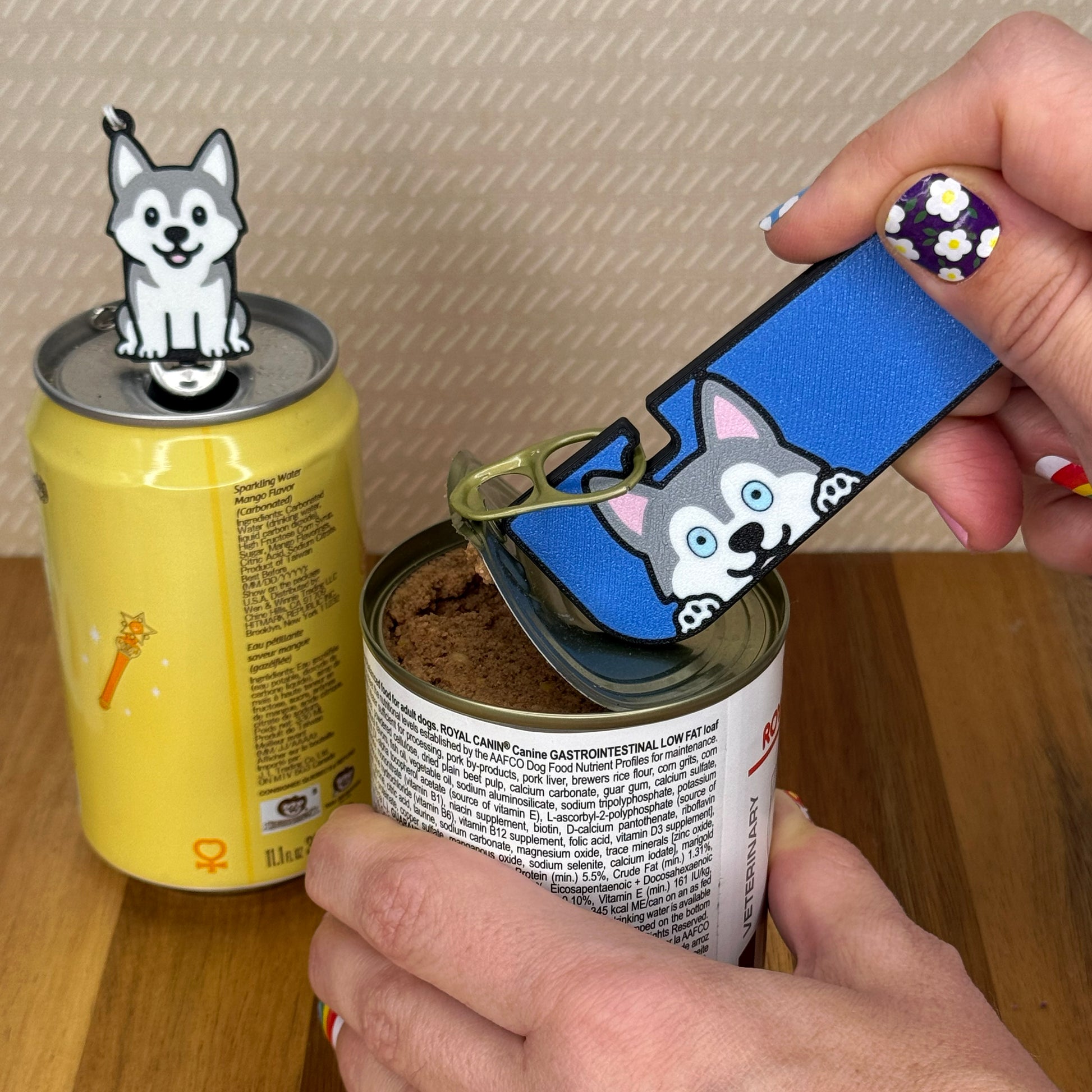 A person opens a can of dog food with a grey Husky dog-design Tab Buddy XL. In the background, a matching soda-sized Tab Buddy rests atop the tab of a can of sparkling water.