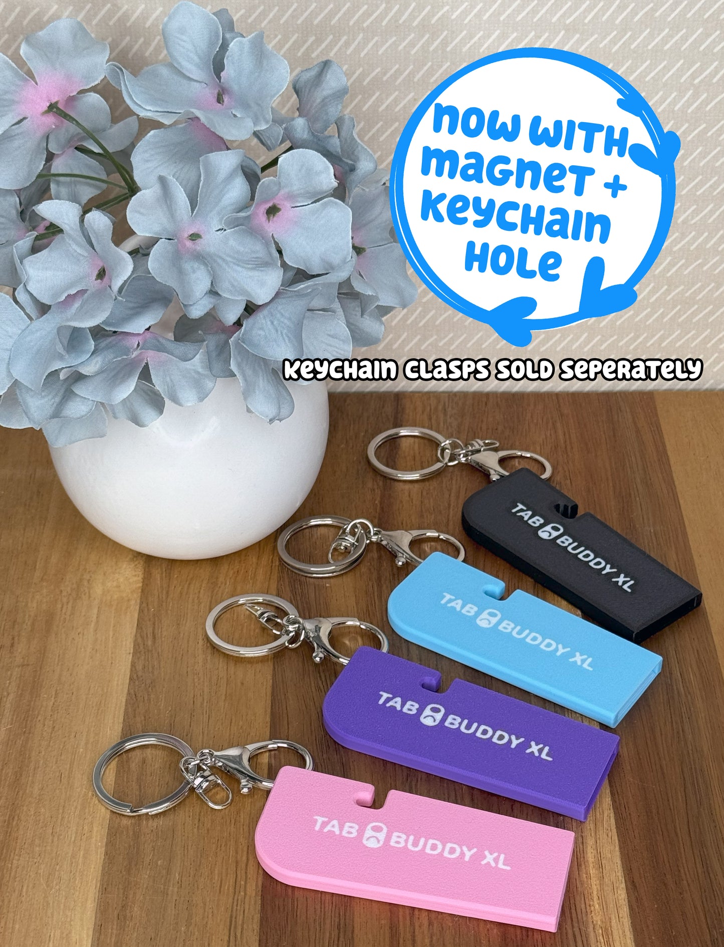 Tab Buddy XLs with attached keychains in various colors on a wooden surface with flowers in the background. Text reads, "Now with magnet + keychain hole; keychain clasps sold seperately."
