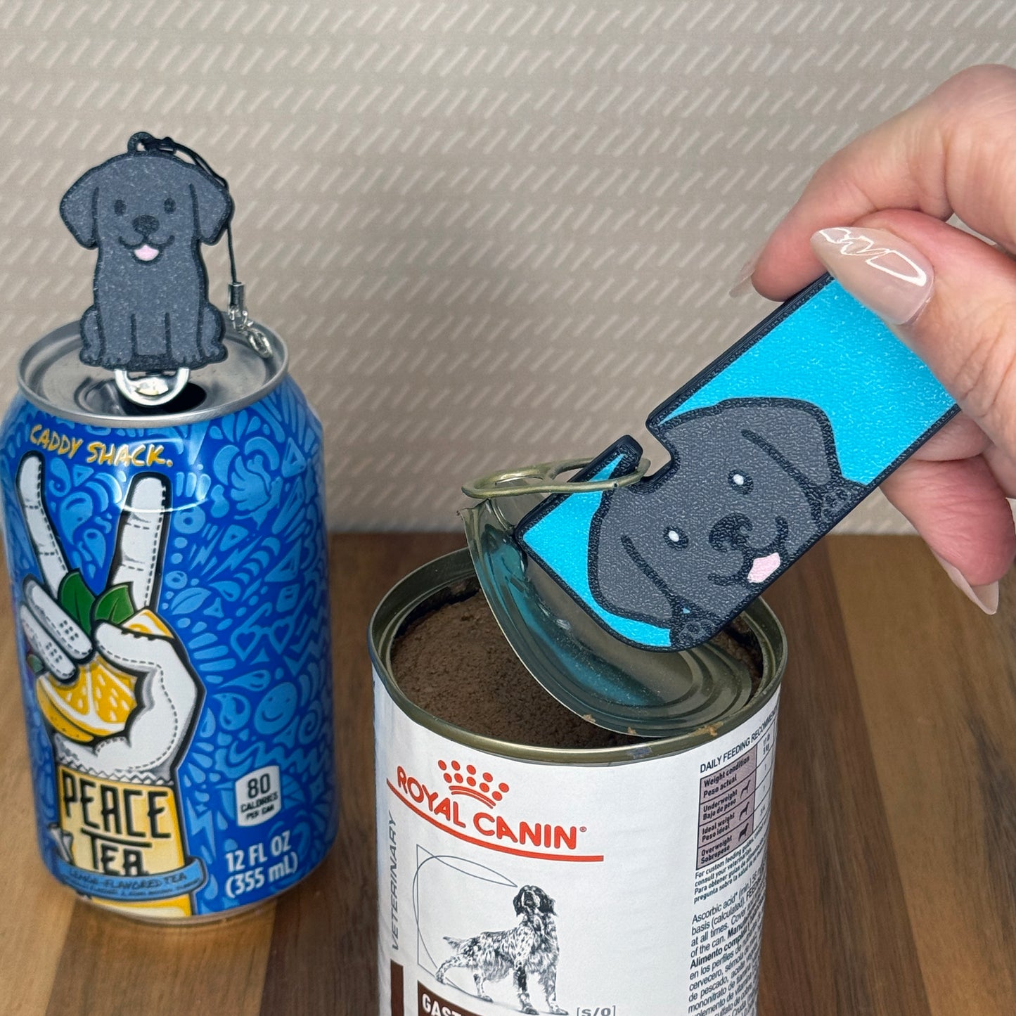 A matching set of Black Labrador-inspired Tab Buddies are shown. The soda-sized Tab Buddy rests atop a blue soda can, and the Tab Buddy XL is used to peel back a can of dog food.