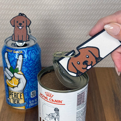 A matching set of Chocolate Labrador-inspired Tab Buddies are shown. The soda-sized Tab Buddy rests atop a blue soda can, and the Tab Buddy XL is used to peel back a can of dog food.