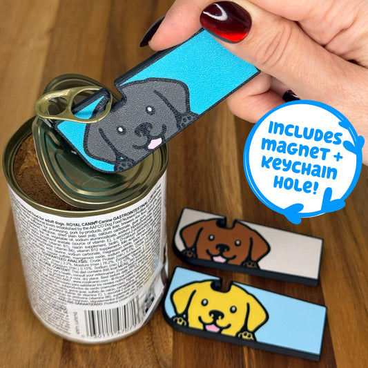 A Black labrador-inspired Tab Buddy XL opens a can of dog food. Two more Tab Buddy XLs in chocolate lab and yellow lab color variations rest on a wooden surface. Descriptive text reads "Includes magnet + keychain hole!"