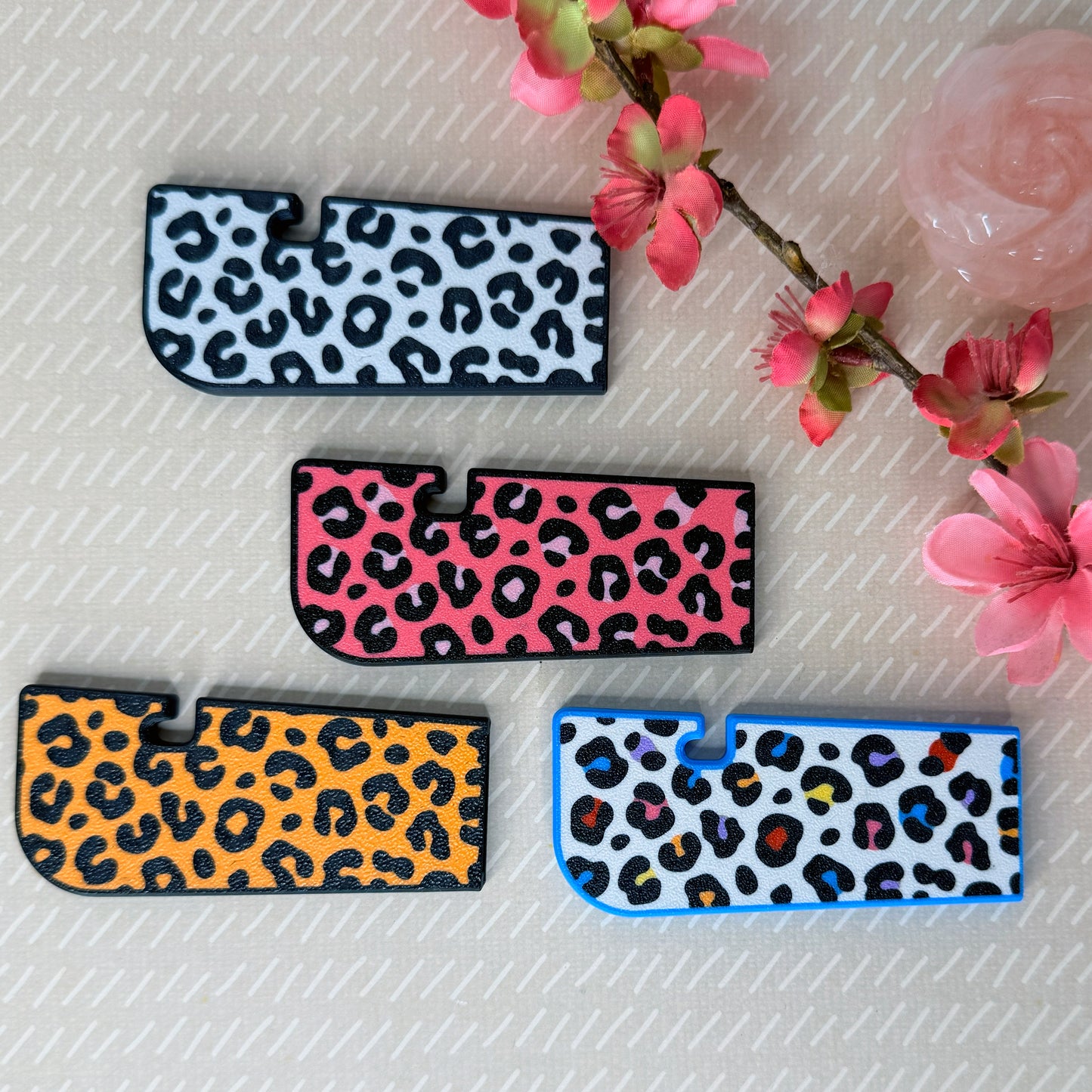Four leopard print Tab Buddy XLs in white, pink, orange and rainbow color vaariations on a light surface with pink flowers.