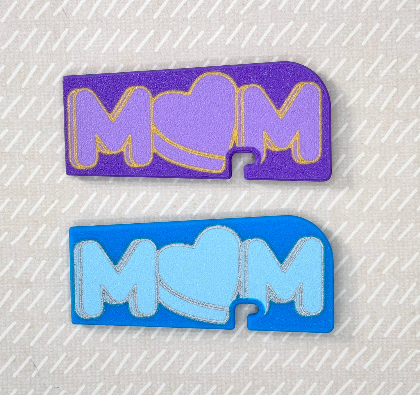 Two 'MOM' Tab Buddy XLs, one purple and one blue, on a textured gray background.