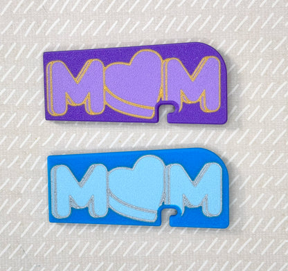Two 'MOM' Tab Buddy XLs, one purple and one blue, on a textured gray background.