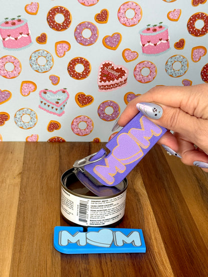 Hand using a Tab Buddy XL with 'MOM' text on a wooden surface and donut-themed wallpaper.