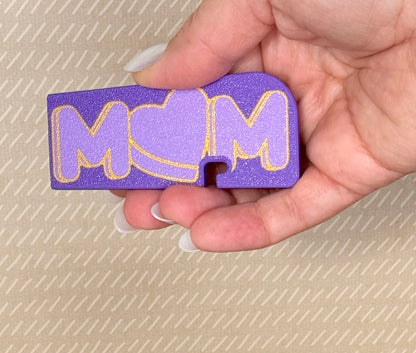 Hand holding a purple 'MOM' Tab Buddy XL with gold accents against a beige background