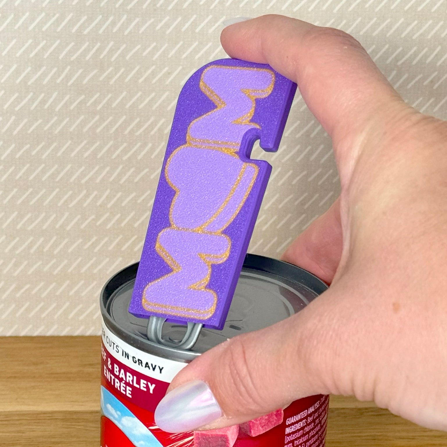 Hand holding a purple 'Mom' Tab Buddy XL to lift the tab on a can of dog food.