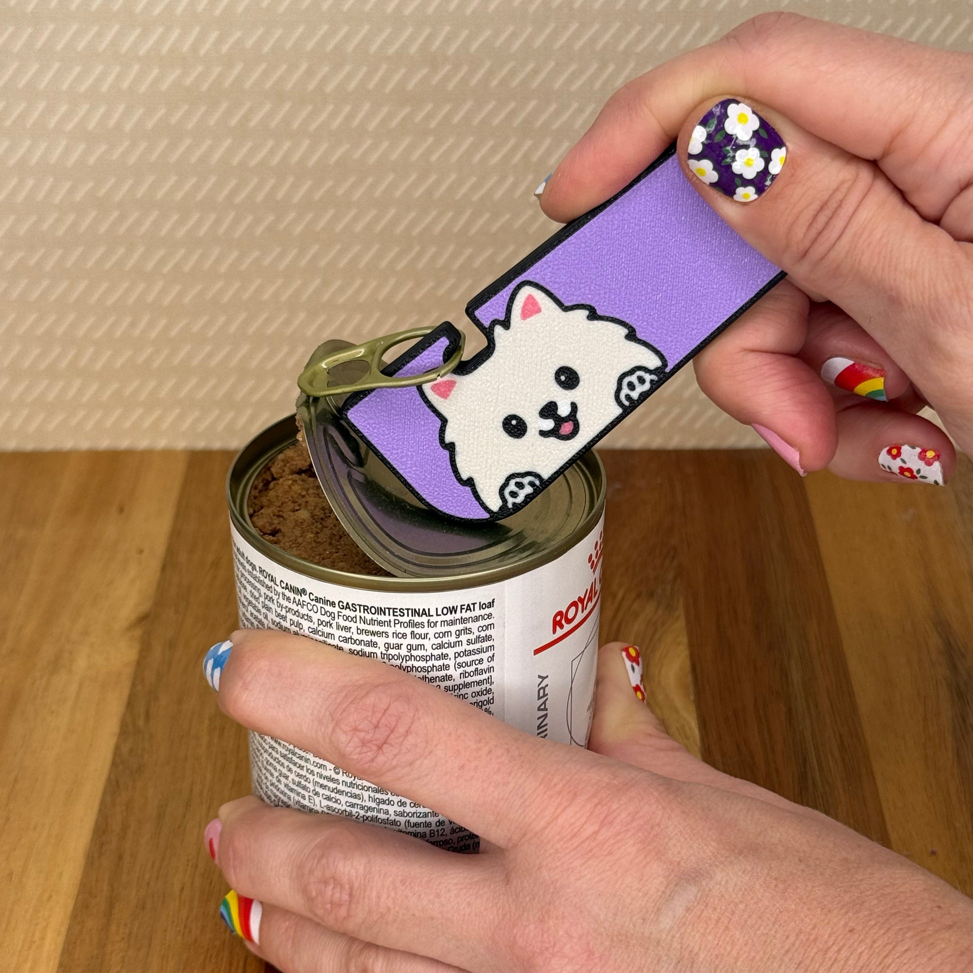 Hand using a Tab Buddy XL with a Pomeranian dog design to open a can of dog food on a wooden surface.