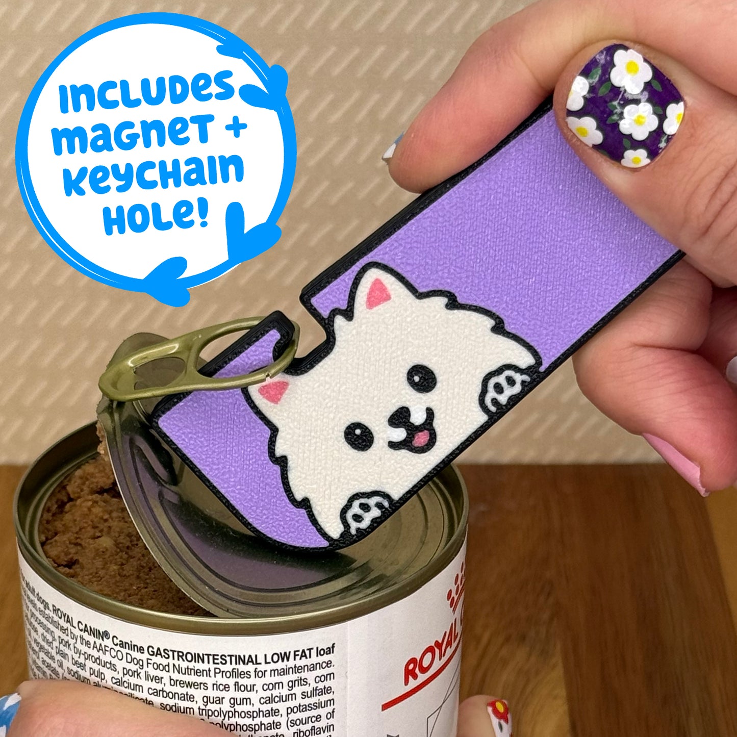Hand using a Tab Buddy XL with a Pomeranian dog design to open a can of dog food on a wooden surface. Text bubble reads, "Includes magnet + keychain strap!"
