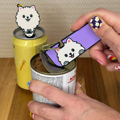 A person opens a can of dog food with a Pomeranian Dog-design Tab Buddy XL. In the background, a matching soda-sized Tab Buddy rests atop the tab of a can of sparkling water.