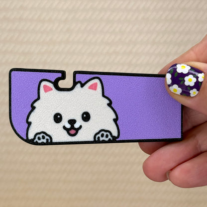 Hand holding a Tab Buddy XL with a cute creamy colored Pomeranian dog design and purple background on its surface. A tan surface is in the background of the photo.