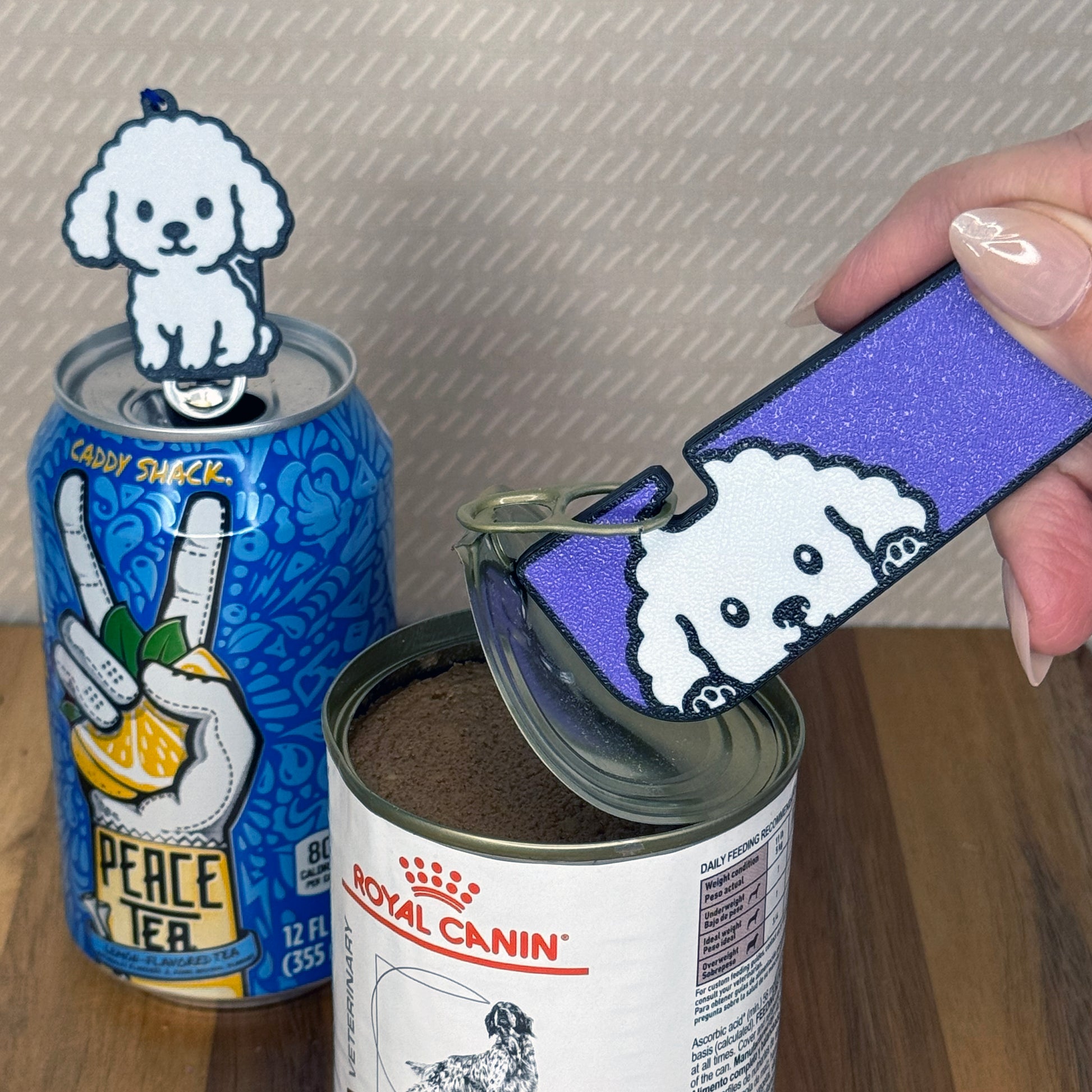 A matching set of white Poodle-inspired Tab Buddies are shown. The soda-sized Tab Buddy rests atop a blue soda can, and the Tab Buddy XL is used to peel back a can of dog food.