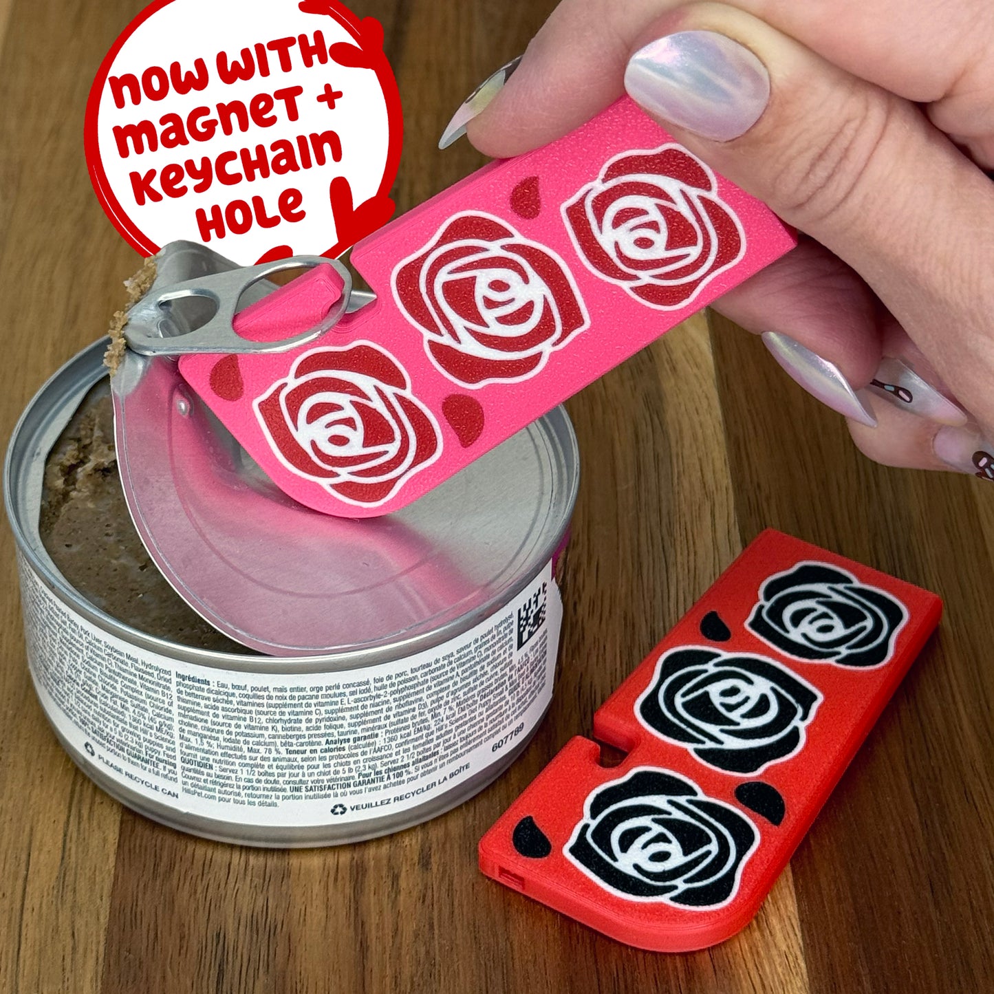 Pink Tab Buddy XL with red rose design being used to open a can on a wooden surface. Another XL, this one red with black roses, rests on the surface. Both come with magnets and keychain holes.