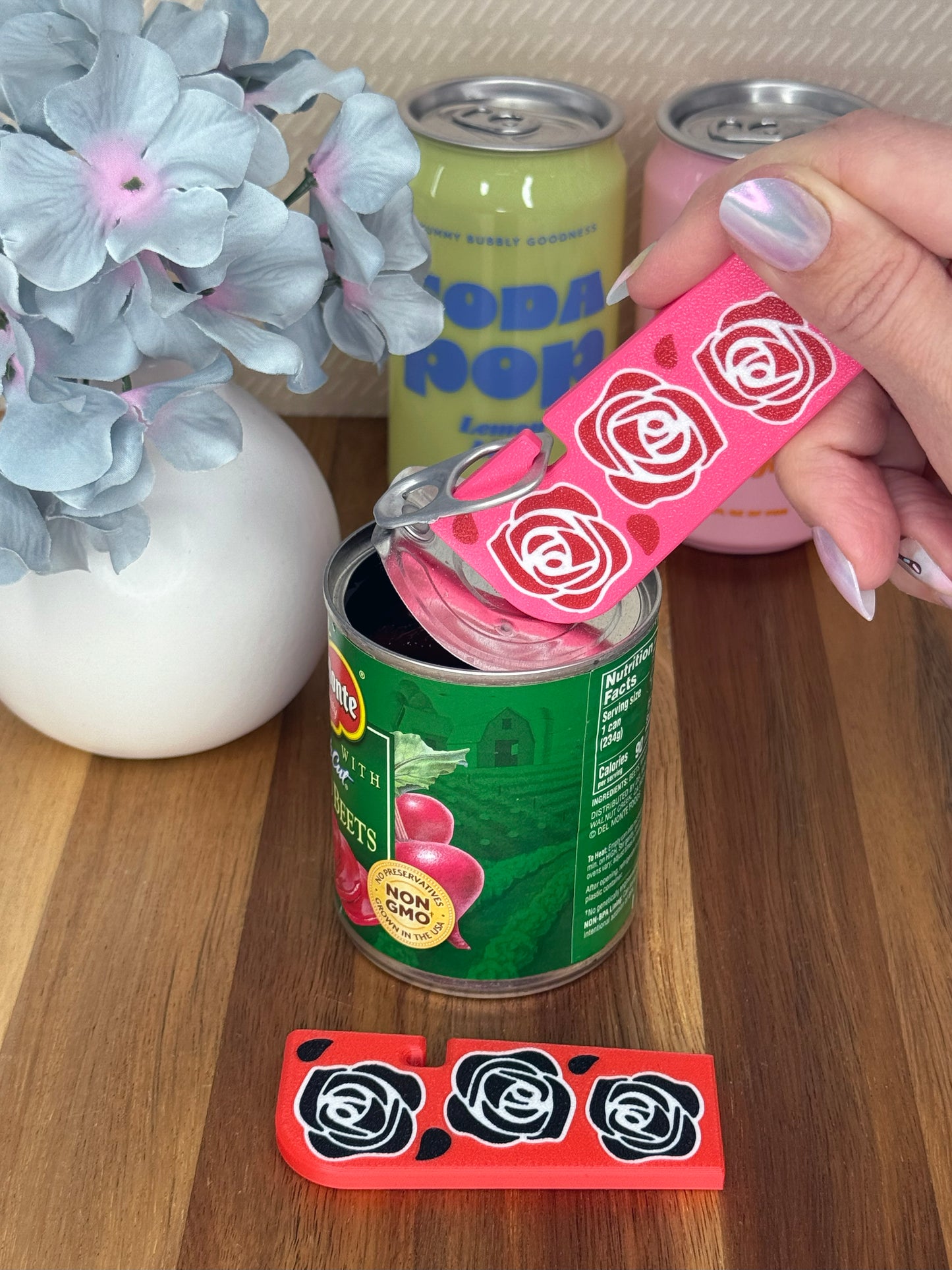 Pink Tab Buddy XL with red rose design being used to open a can on a wooden surface. Another XL, this one red with black roses, rests on the surface. Flowers and soda cans decorate the background. 