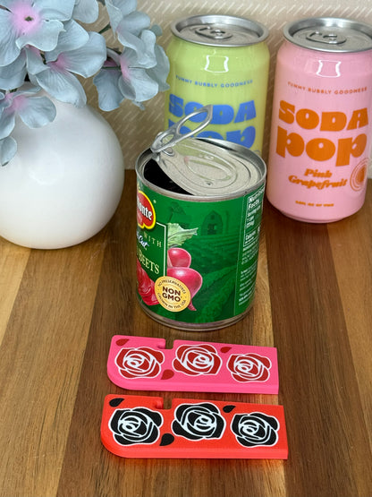 Two Tab Buddy XLs -- one pink with red rose design and one red with black roses, rest on a wooden surface in front of an open can of beets.