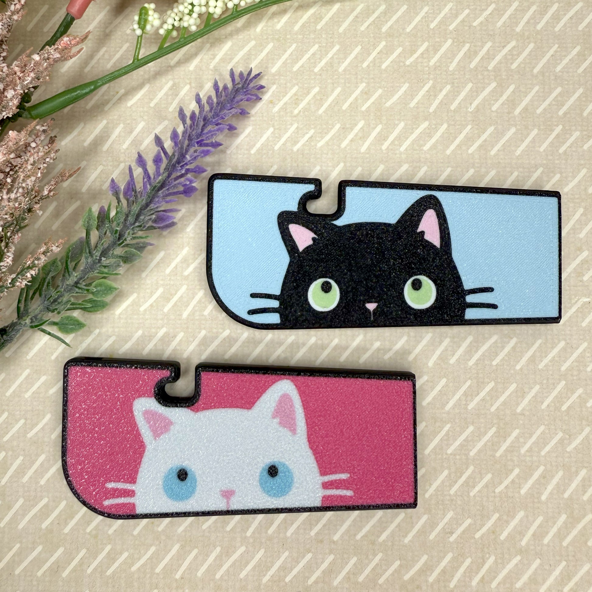 Two cat-themed Tab Buddy XL, one black and one white, on a textured beige background with flowers.