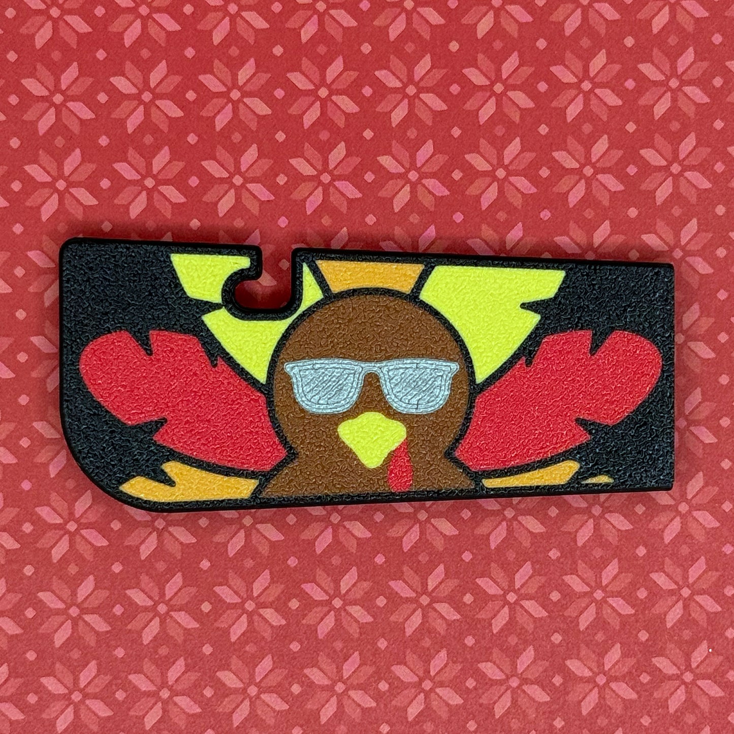 Tab Buddy XL with black backgrou7nd and red, orange and yellow feathers wearing silver sunglasses rests on a red floral patterned background