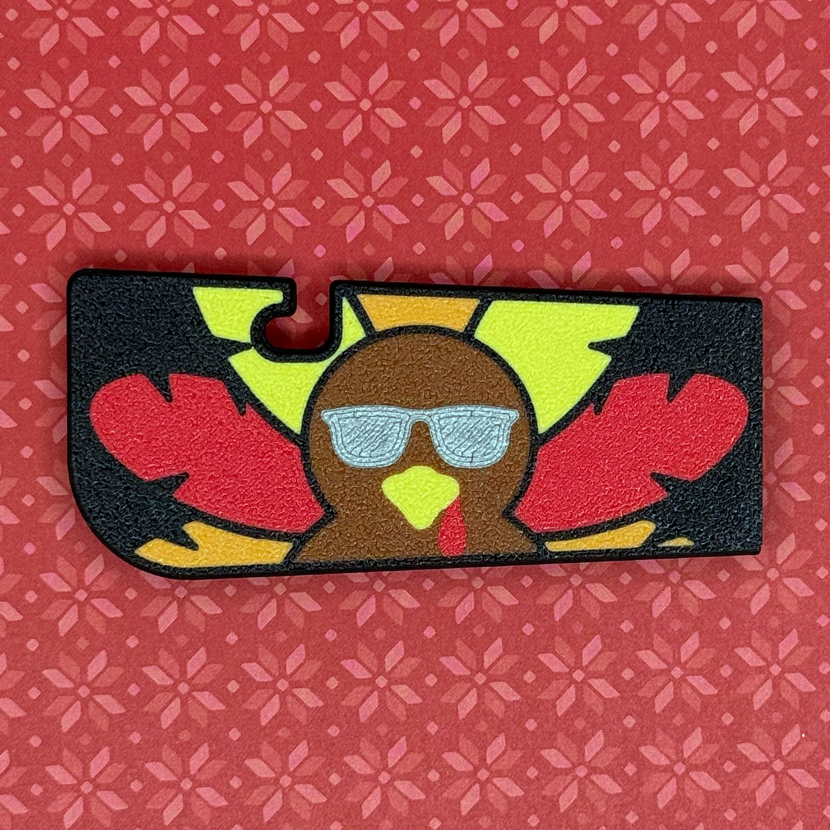 Tab Buddy XL with black backgrou7nd and red, orange and yellow feathers wearing silver sunglasses rests on a red floral patterned background