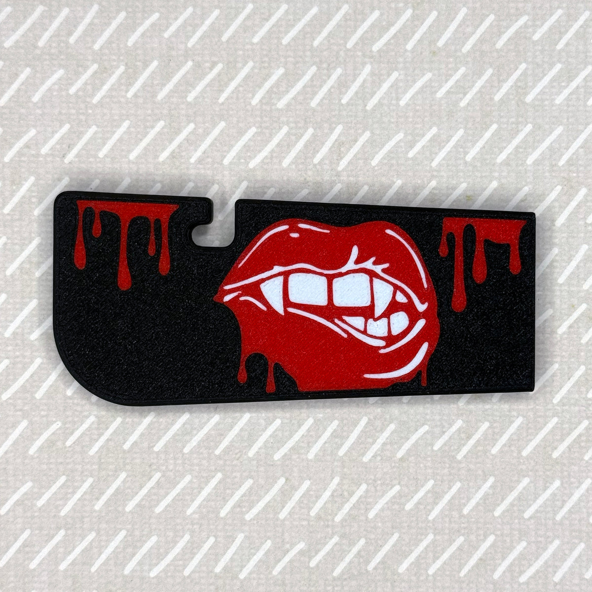 A black Vampire Lips Tab Buddy XL rests against a beige patterned backgeound.