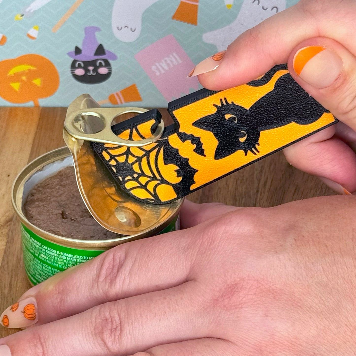 Hand using a Halloween-themed black cat Tab Buddy XL on a cat food can with a colorful background featuring pumpkins and ghosts.