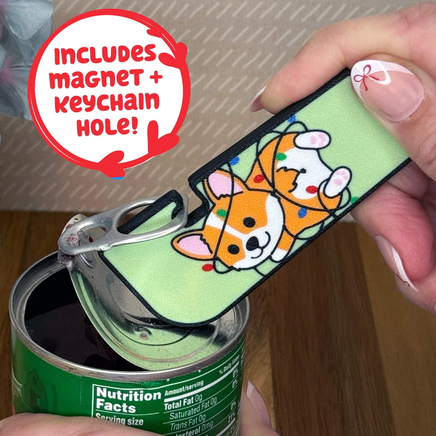 Person using a Tab Buddy XLwith a cartoon style corgi wrapped in Christmas lights on a green background is used to open  a can of vegetables. Text notes the Tab Buddy XL includes a magnet and keychain hole.