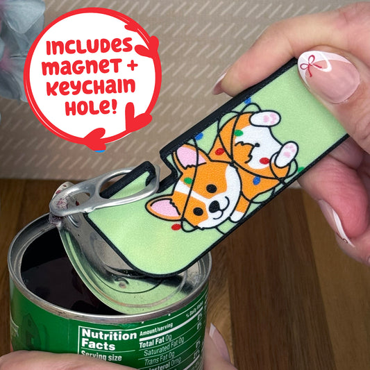 Person using a Tab Buddy XLwith a cartoon style corgi wrapped in Christmas lights on a green background is used to open  a can of vegetables. Text notes the Tab Buddy XL includes a magnet and keychain hole.