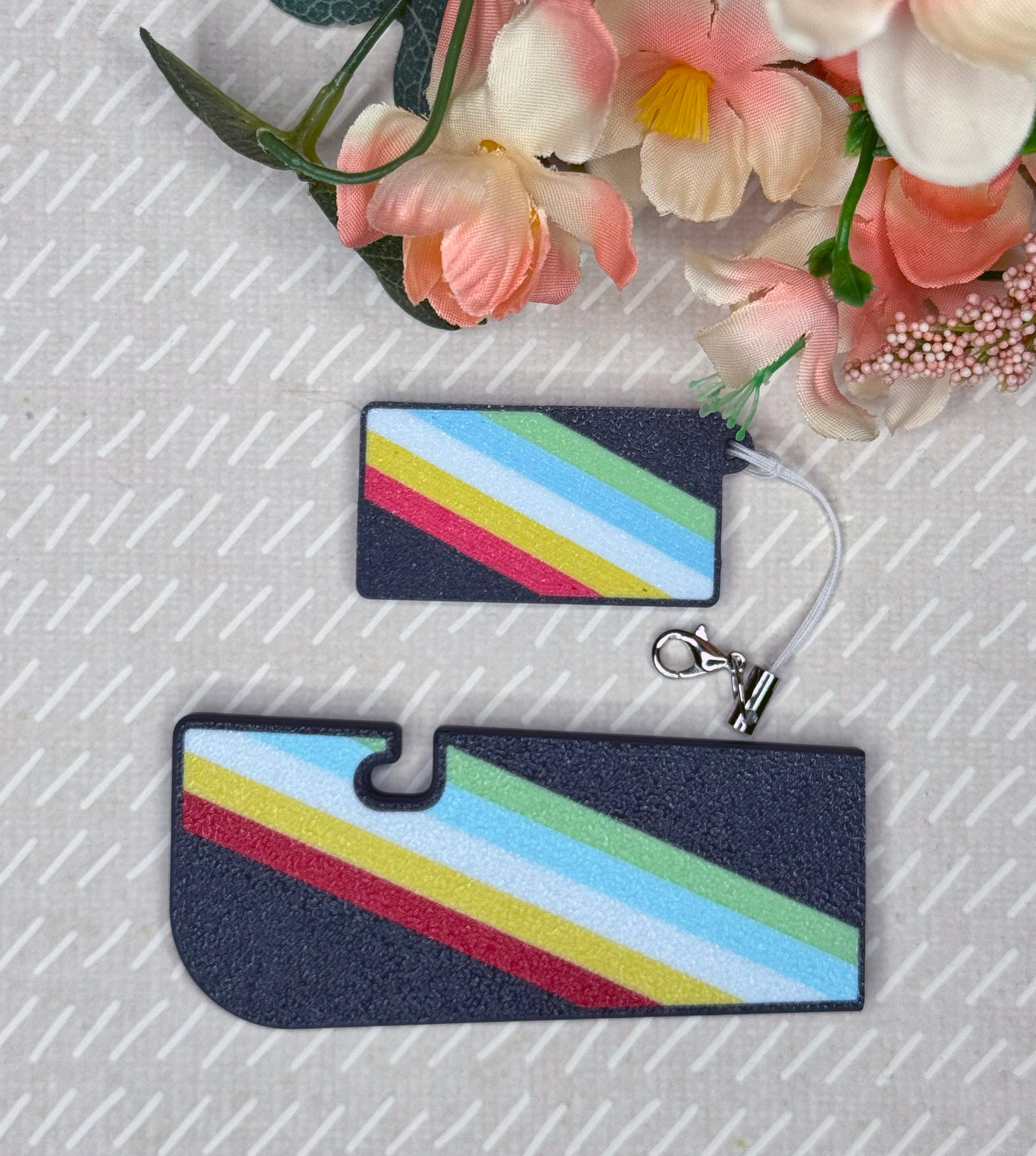 Matching set of disability pride Tab Buddies -- one soda Tab Buddy and one Tab Buddy XL -- on a textured surface with flowers in the background