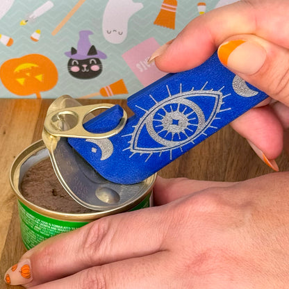 Hand using a blue Tab Buddy XL with a decorative design on a can against a Halloween-themed background.