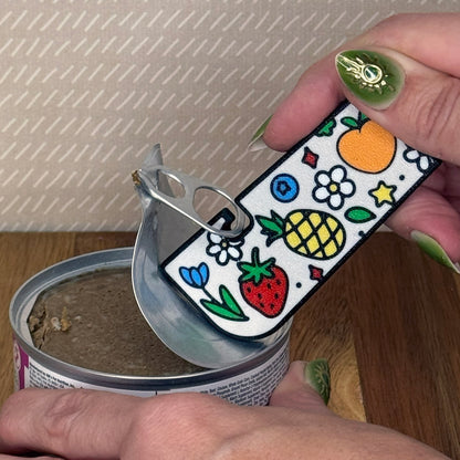A Tab Buddy XL with a colorful Fresh and Fruity design is used to peel back the tab lid on a can of cat food. The design includes a peach, pineapple, strawberry, blueberry, and flowers.
