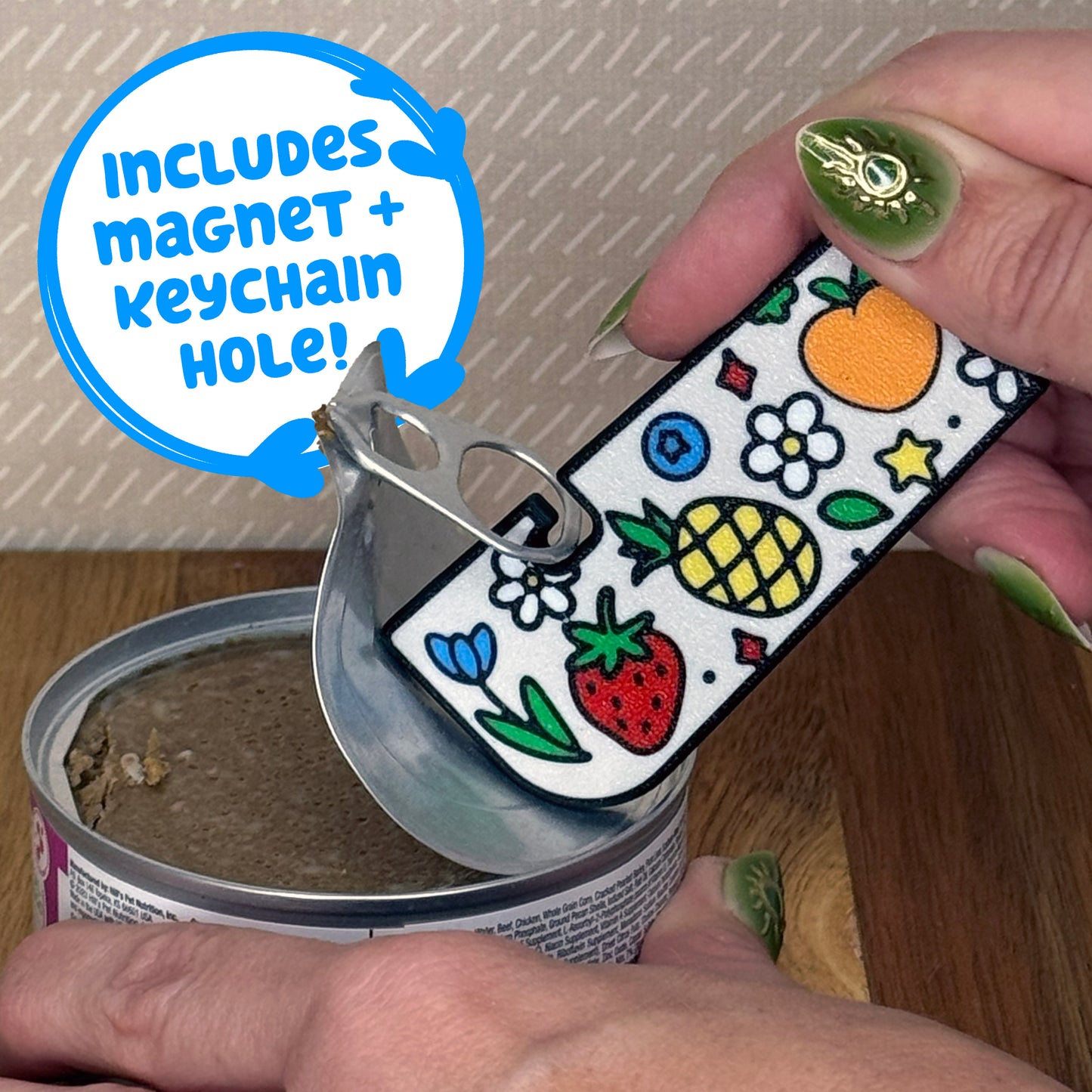 A Tab Buddy XL with a colorful Fresh and Fruity design with flowers and fruit on it is used to peel back the tab lid on a can of cat food. Text reads "Includes magnet + keychain hole!"