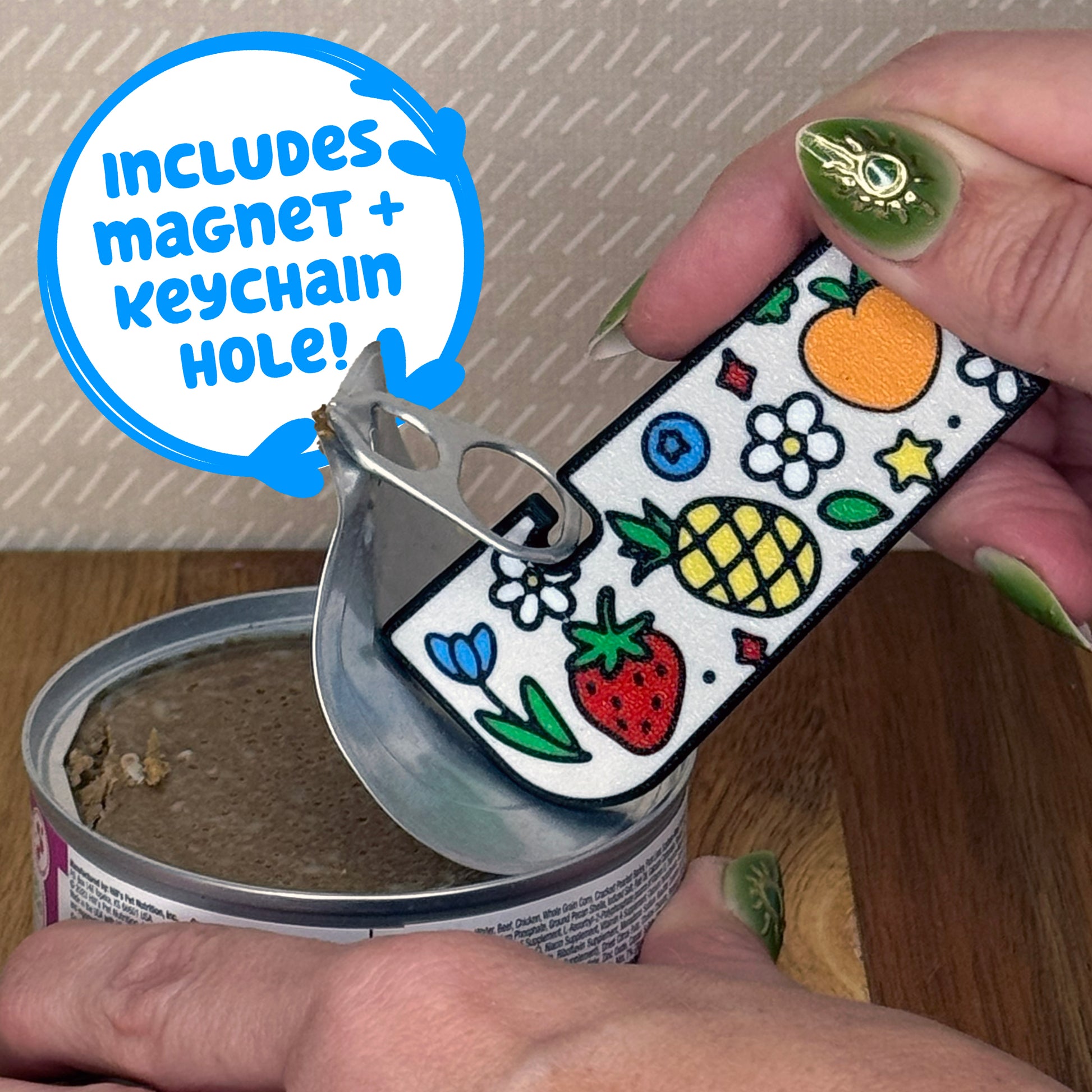 A Tab Buddy XL with a colorful Fresh and Fruity design with flowers and fruit on it is used to peel back the tab lid on a can of cat food. Text reads "Includes magnet + keychain hole!"