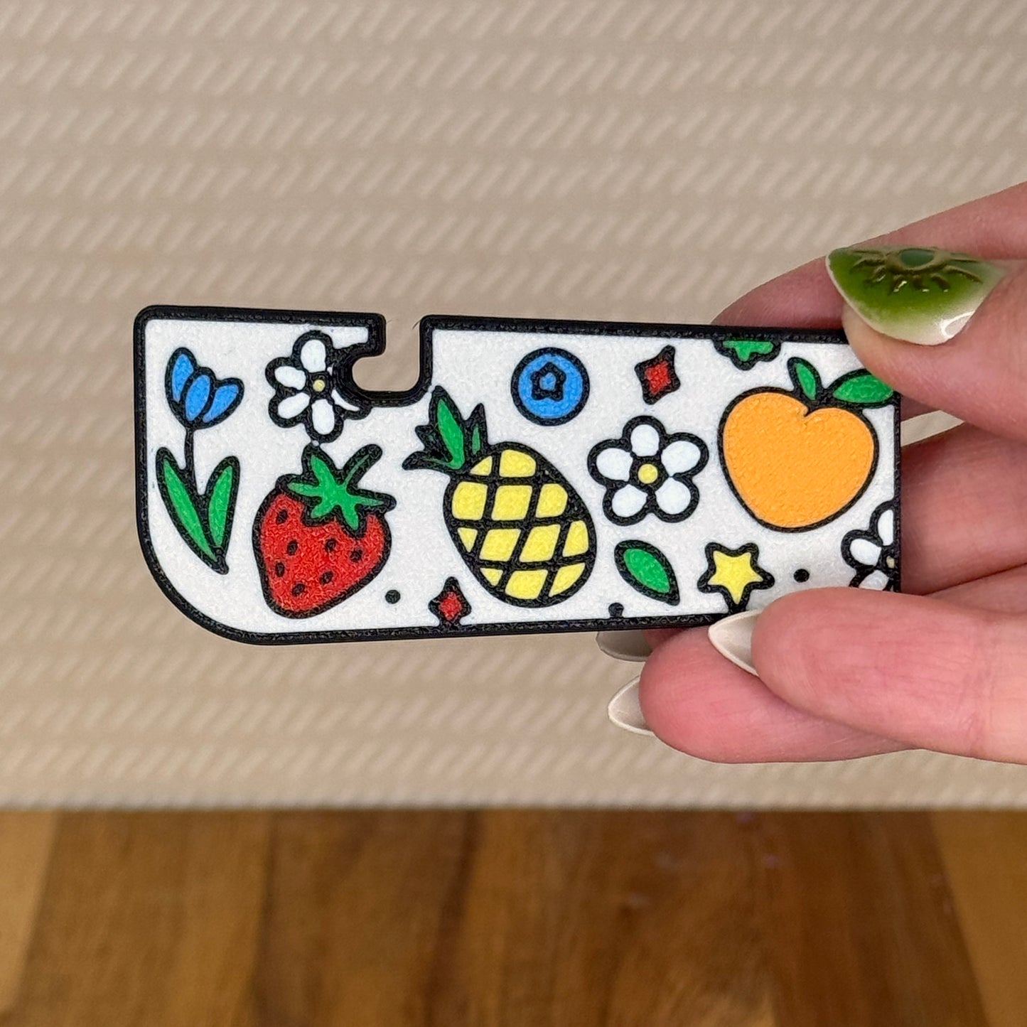 A Tab Buddy XL with a colorful Fresh and Fruity design is held in a hand against a beige background. The design includes a peach, pineapple, strawberry, blueberry, and flowers.