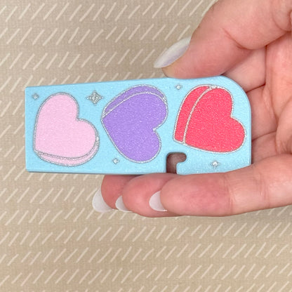 Hand holding a blue Tab Buddy XL with three colorful hearts on a beige background