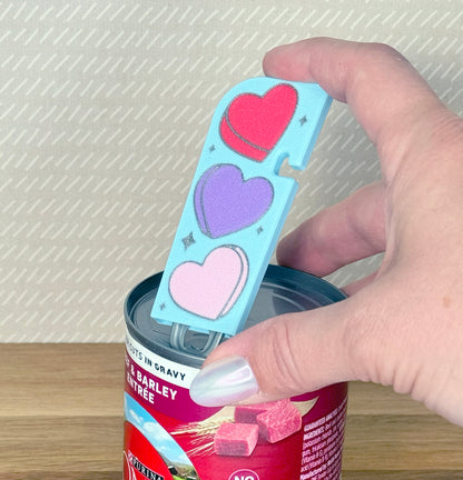 Hand using a blue Tab Buddy XL with heart designs to lift the tab on a can of food.